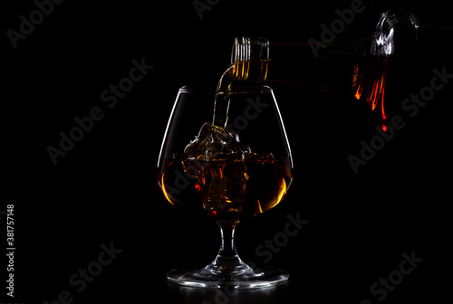 Pouring whiskey drink into glass with ice cubes on black background