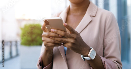Close up of smartphone in hands of African American young businesswoman while tapping and scrolling on screen. Woman texting message on mobile phone outdoor. Female using gadget.