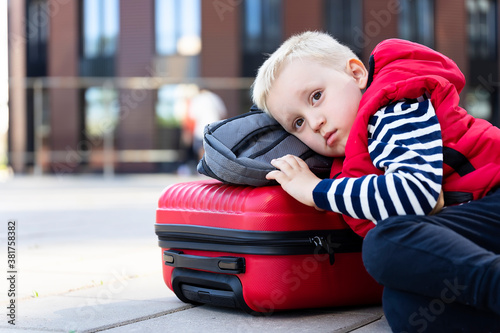 Tired little boy traveler on the background of modern buildings lies on a suitcase