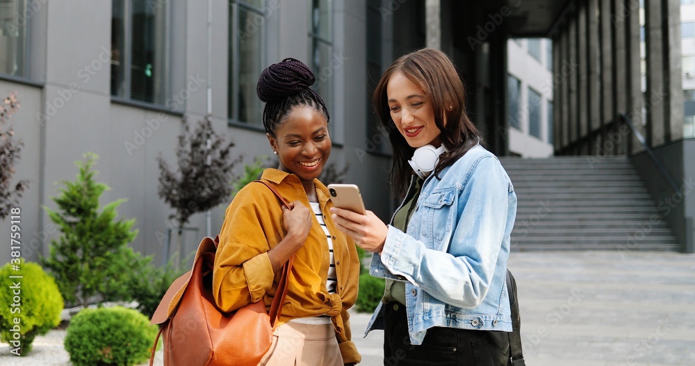 Mixed-races females talking and watching something on mobile phone at street in city. Beautiful multi ethnic young women chatting and using smartphone. Cheerful friends gossiping. Gossips.