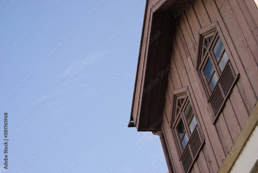 Topo de uma casa construído em madeira com duas janelas e o céu a fazer fundo, formas triangulares cores azuis e castanhas