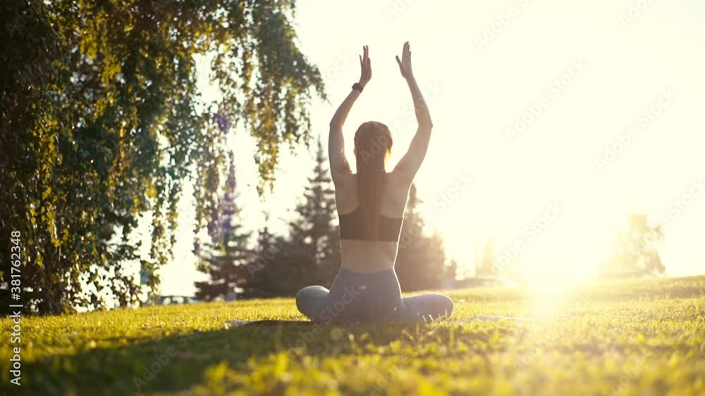 Back view of unrecognizable slender young woman sitting on yoga mat in lotus position and raising hands up outside in city park. Rear view of female practicing yoga outdoors in sunny day.