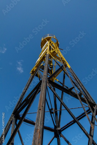 a photograph of an old somewhat rusty lighthouse painted black and yellow with a blue sky background and vertically cropped