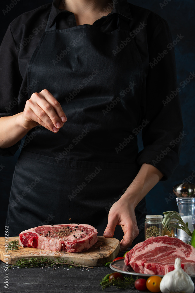 The chef in black apron pours pepper on the fresh beef steak on dark background. Backstage of preparing traditional restaurant or hotel meal. Food concept. Frozen motion. Adds spices to the dish.