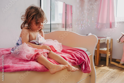 little upset girl sits on the bed in her room and holds a dress. she can't choose clothes