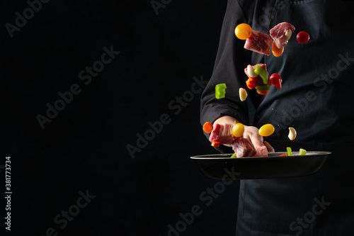 The chef in black apron throwing the fresh meat, beef or pork, mixed with vegetables, on dark background. Frozen motion. Food concept.
