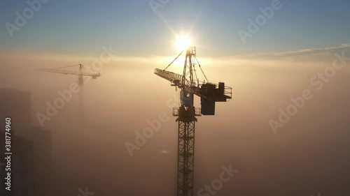 Construction site at dawn. Tower cranes over fog on the background of the morning sun. Drone view
