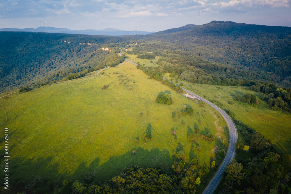 Fototapeta premium Early morning in the mountains. Morning sun and haze in the valley against the background of mountains. Green fields and meadows. The road leaving into the distance. Adygea, Russia