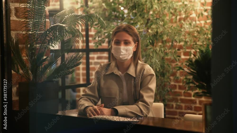 Young woman with protective mask sits in cafe near window and waves hand to her friend outside Young woman with protective mask sits in cafe near window and waves hand to her friend outside