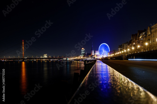 Night scenery over fence on promenade along riverside of Rhine river and background of Ferris wheel at Christmas market and cityscape in D  sseldorf  Germany.