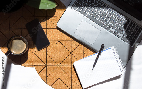 Vista cenital de un escritorio moderno con una plancha de corcho de figuras geométricas bajo la luz del sol de la mañana. Oficina en el hogar con laptop, teléfono móvil, anotador y una taza de café.