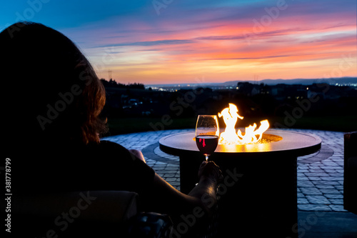 Fototapeta A woman relaxes with a glass of wine at sunset by a fire pit on the patio of a l