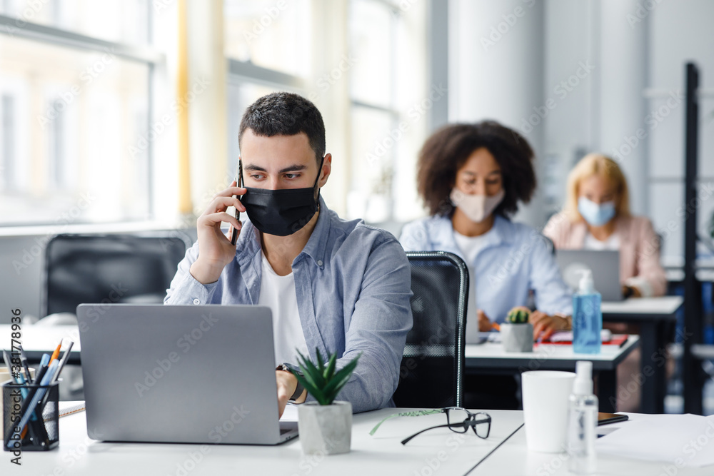 Customer consultation by phone remotely at returning to work after quarantine. Millennial man in protective mask with smartphone looks at laptop at workplace Customer consultation by phone remotely at returning to work after quarantine. Millennial man in protective mask with smartphone looks at laptop at workplace