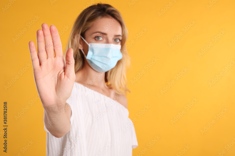 Woman in protective face mask showing stop gesture on yellow background, focus on hand. Prevent spreading of coronavirus
