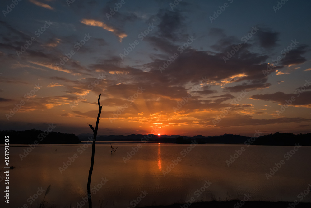 Reservoir or lake in the evening with dry trees in the middle.The sun is falling behind the clouds and there are beams of light coming out.