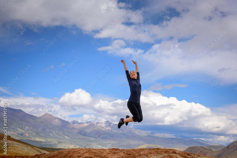 Young woman enjoying life and jumping against blue sky and mountains background. Happy female tourist jumps with hands up on top of rock on summer sunny day. Travel, freedom concept. Young woman enjoying life and jumping against blue sky and mountains background. Happy female tourist jumps with hands up on top of rock on summer sunny day. Travel, freedom concept.