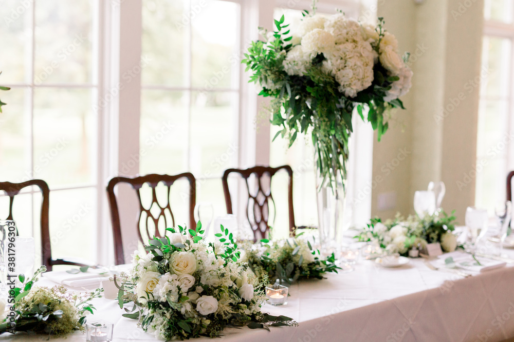 The head table at a wedding reception with the bride and bridesmaids bouquets on the table. The table is decorated with tall standing glass vase with a huge white and green floral arrangement. 