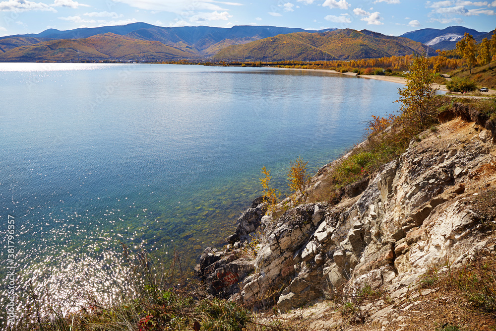 Autumn landscape with beautiful yellowed trees on the shore of lake Baikal on a sunny day. Calm clear water, pebble beach.