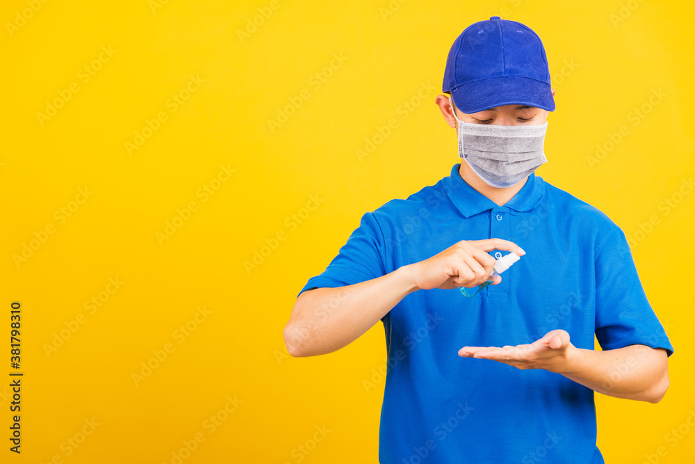 Asian young handsome delivery man wearing face mask protective germ virus and mask alcohol sanitizing sprays to hand, studio shot isolated on yellow background, medical outbreak coronavirus COVID-19 Asian young handsome delivery man wearing face mask protective germ virus and mask alcohol sanitizing sprays to hand, studio shot isolated on yellow background, medical outbreak coronavirus COVID-19