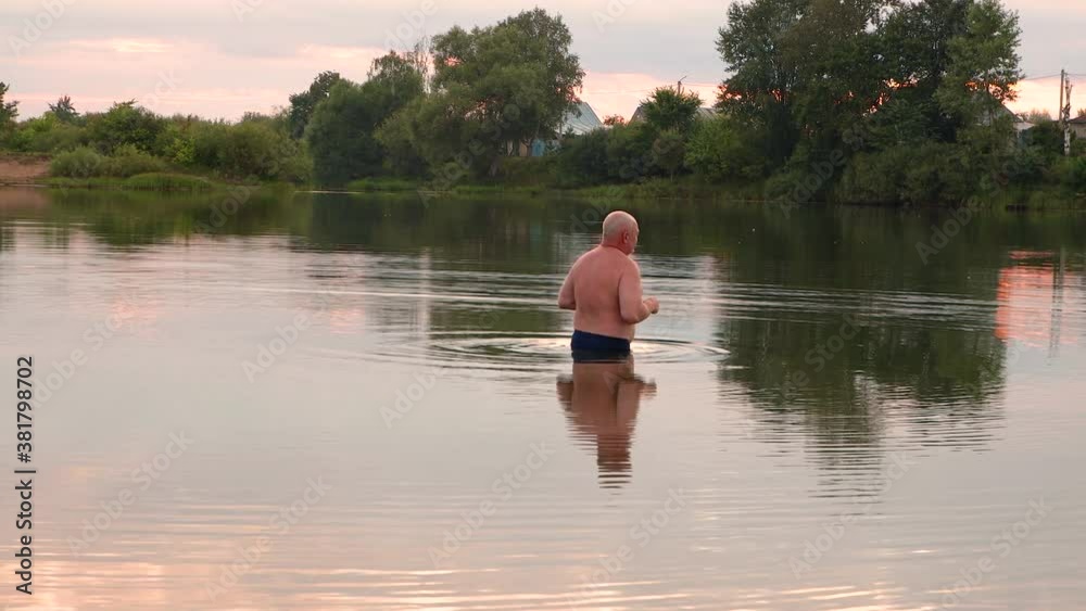 A man enters the cool pond and dives. Cloudy sky and green trees are reflected in the lake. The sun gives off a reddish light at sunset. Summer evening.