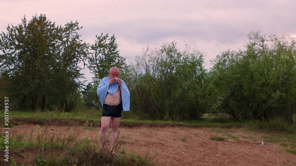 A man in swimming trunks is standing on the green grass. There is a blue towel on the shoulders. The man is drinking a drink. Against the background of a red gloomy sky. Evening time of the day. A man in swimming trunks is standing on the green grass. There is a blue towel on the shoulders. The man is drinking a drink. Against the background of a red gloomy sky. Evening time of the day.