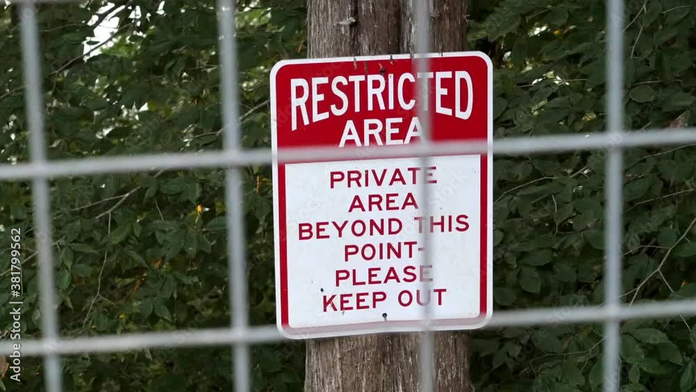 Sign: Restricted Area, Private area beyond this point, please keep out. Red and white warning sign viewed through open metal fence, trees in background. Sign: Restricted Area, Private area beyond this point, please keep out. Red and white warning sign viewed through open metal fence, trees in background.