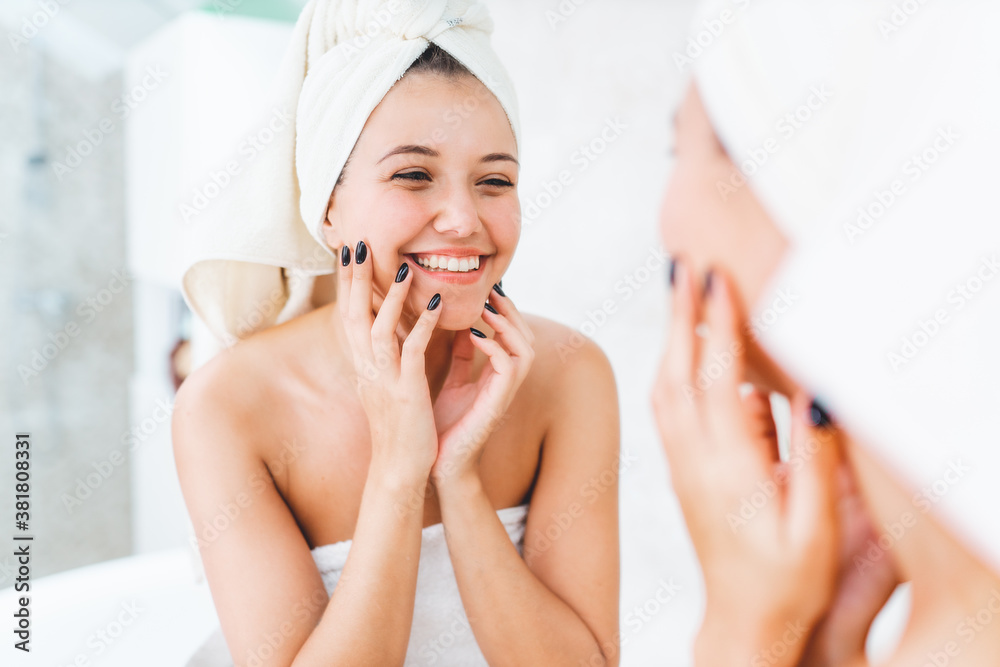Young attractive woman with white towel on head and body with reflection in mirror in bathroom. Female smile and look at herself Young attractive woman with white towel on head and body with reflection in mirror in bathroom. Female smile and look at herself