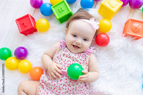 a beautiful little baby girl is lying in red clothes on a white Mat among toys balls and cubes