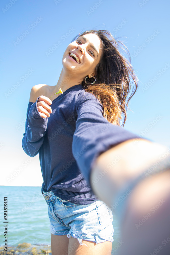 Vertical shot, selfie of young brunette italian woman with long hair in dark blue sweater, romantic with hair in the wind happy, smiling and with intense blue sky and with a flower in her right hand