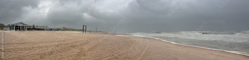 A severe windstorm hits the beach in summer. Under a dramatic sky the wind creates a fierce surf and blowing sand hits the deserted beach pavilions. A severe windstorm hits the beach in summer. Under a dramatic sky the wind creates a fierce surf and blowing sand hits the deserted beach pavilions.