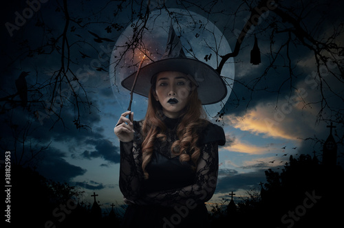 Halloween witch holding magic wand standing over cross, church, crow, bat, birds, dead tree, full moon and sunset sky, Halloween mystery concept photo