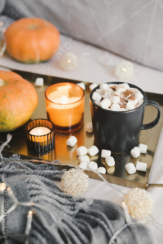 Hot chocolate with melting mini marshmallows served in bed on a metal tray with pumpkin, burning candles and decorative lights. Autumn season inspiration