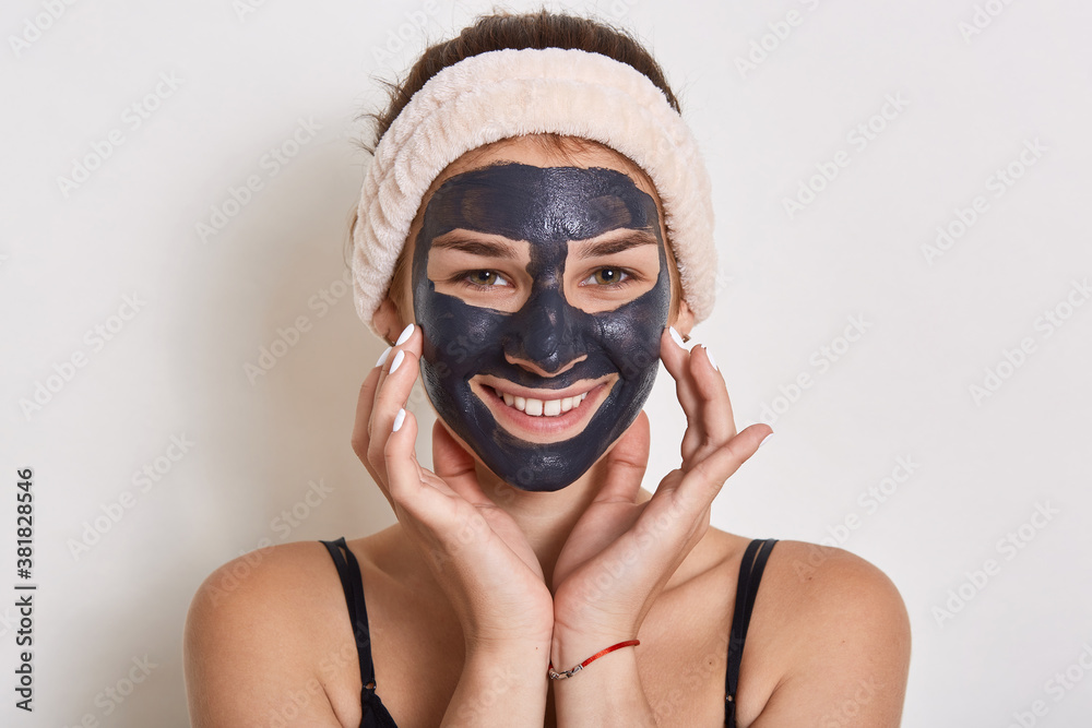 Smiling lady posing isolated over white background, girl in hair band posing with black facial mask, looks at camera, beauty treatment in morning at home. Smiling lady posing isolated over white background, girl in hair band posing with black facial mask, looks at camera, beauty treatment in morning at home.