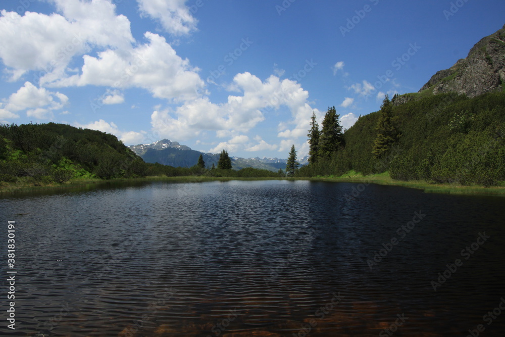 Blauer Bergsee in den Alpen mit Wiese und Wald an einem sonnigen Sommertag