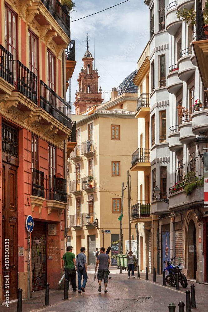Valencia, Spain: beautifully restored alley in the old town; in the back a bell tower; people stroll along the street