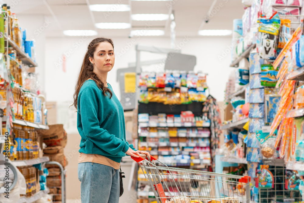 Portrait of a young pretty Caucasian woman in casual clothing poses with a grocery cart in the aisle of a store. Copy space. The concept of shopping and buying food