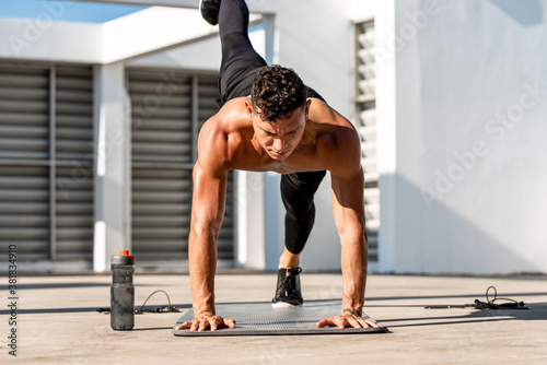 Shirtless muscular male athlete doing high plank leg lift exercise in the open air on building rooftop floor