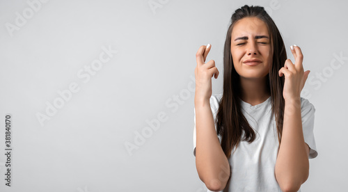 Young woman raises fingers crossed, makes desirable wish, waits for good news. Copy space. Studio shot white background © AstiMak