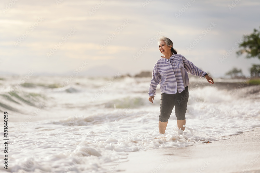 Senior Asia woman dancing on the beach with barefoot, Smile with Happily while wave in the sea rush to shore