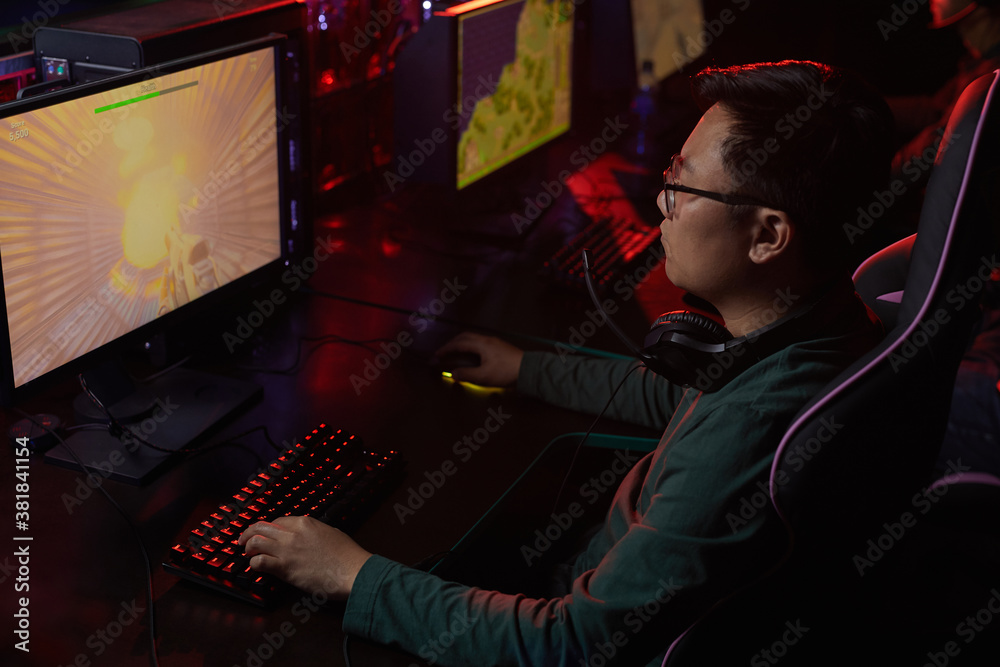 Asian young gamer in eyeglasses looking at computer monitor he concentrating on his game in dark computer club Asian young gamer in eyeglasses looking at computer monitor he concentrating on his game in dark computer club