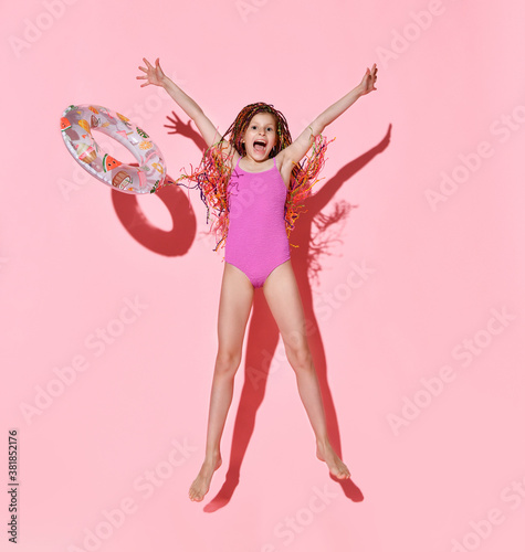 Happy girl with bright cornrows in one-piece rose swimsuit jumping and screaming full of joy, throwing rubber ring with fruit pattern
