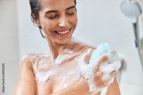 Cheerful young woman using exfoliating loofah while taking shower