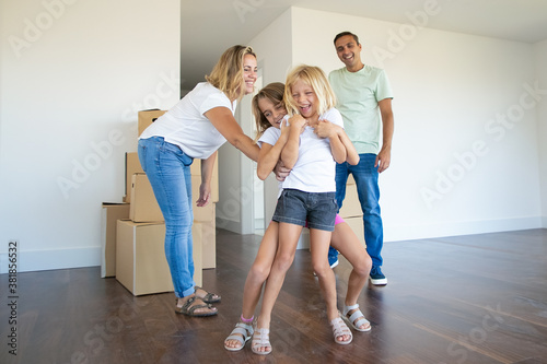Joyful family couple and two kids having fun while moving into new apartment. Girls tickling each other and laughing. Full length. Real estate purchase concept