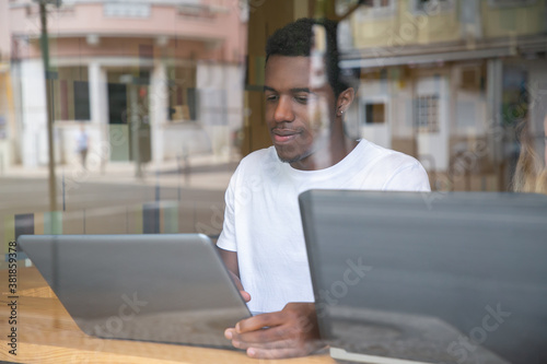 Content African American guy sitting at table and using laptop. Attractive concentrated man behind window working on portable computer indoors. Freelance and digital technology concept