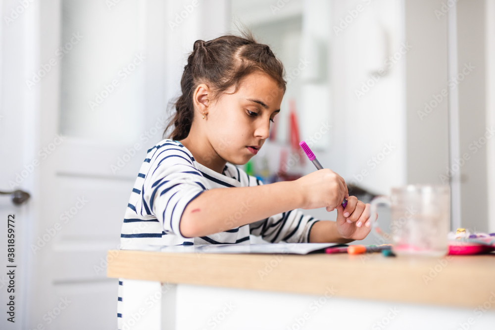 Cropped shot of a focused little girl making a picture using different types of art supplies while being seated at a table inside during the day Cropped shot of a focused little girl making a picture using different types of art supplies while being seated at a table inside during the day