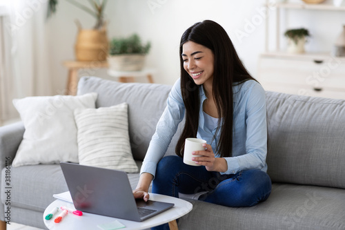 Positive Asian Freelancer Woman Working On Laptop And Drinking Coffee At Home