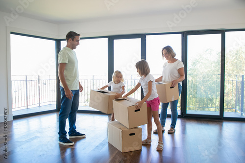 Smiling mother with kids holding cardboard boxes and putting them in stack. Caucasian father standing, looking at girls and talking. Home owners watching apartment. Relocation and moving day concept