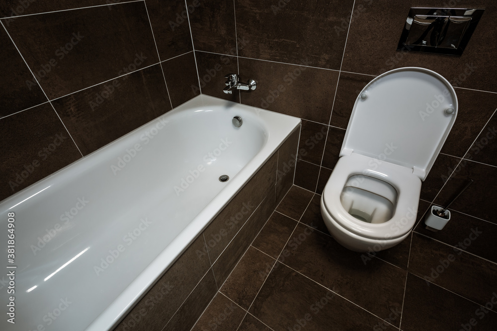 Fototapeta premium Detail of a small bathroom with a bathtub and a toilet next to each other. The toilet is modern with an open seat. Paving and tiling have the texture of a concrete trowel in shades of brown.