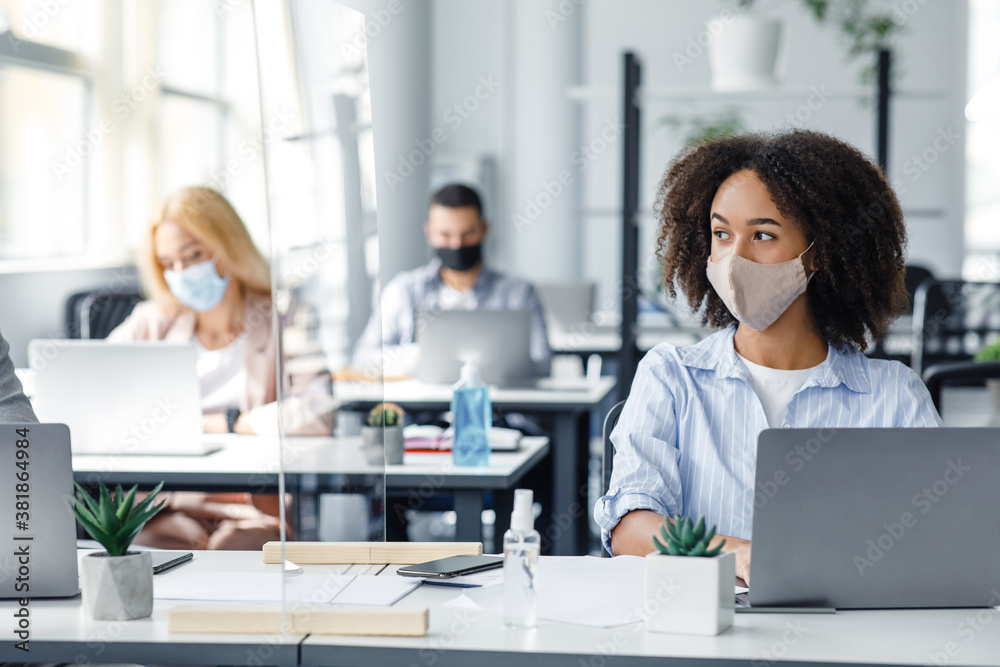 Work on project in corporate company after lockdown. African american woman in mask creating idea at workplace with smartphone and protective glass Work on project in corporate company after lockdown. African american woman in mask creating idea at workplace with smartphone and protective glass