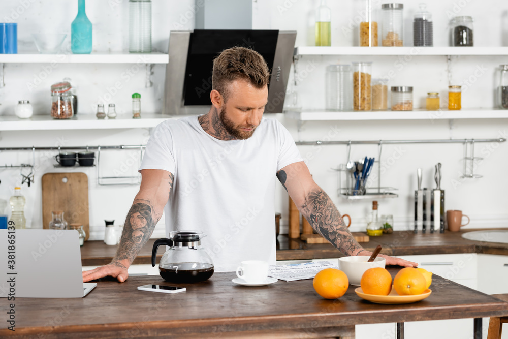 young man in white t-shirt reading newspaper during breakfast near laptop in kitchen